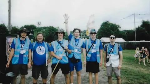 A film photo showing a group of six WaterAid volunteers wearing hi-vis branded blue vests and lanyards. They are carrying equipment like a mop and a bucket to clean toilets. They are smiling and posing for a photo.