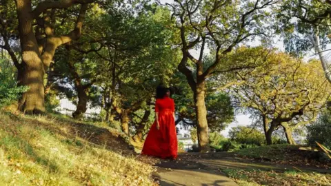 Amaal Said A black woman walking through a sunny park, filled with trees, in a bright red dress.