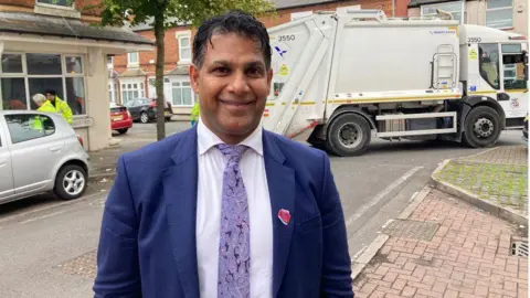 A man with short black hair, wearing a blue suit jacket, a white shirt and purple patterned tie, stands smiling in a street. A refuse collection vehicle is pulling out of a junction behind him.