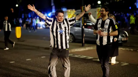 Two fans crossing the street wearing black and white Newcastle tops. It is dark and one of the men is smiling and is putting his hands out. 