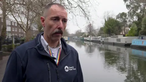 A man stands at the side of a canal with houseboats in the background