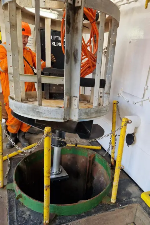 UKCSM A man in orange overalls and hard hat is standing near a round metal cage which is moving through the ship's floor through a round hole towards the sea. The hole has four poles and chain around it.