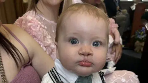 A baby, Eli, pictured in a church. He is wearing a white skirt, a blue bow tie and black suspenders. He has strawberry blonde hair and is sitting on a woman's lap, she is wearing a pink spaghetti strap dress. The woman next to them is wearing a pink dress.