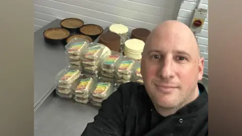 Jonny Catling A selfie of Jonny Catling in the bakery's kitchen. He is bald and wearing black chef clothes. He is standing in front of rows of cakes, which are ready for sale.