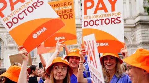 Junior doctors protest in August 2023, wearing blue scrubs and orange hats hold placards reading: "Pay restoration for doctors".