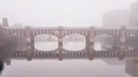 David Pollok The  Caledonian Railway Bridge over the River Clyde in Glasgow. The bridge has red and white metal connected between red brick turrets. A thick fog is covering the scene and the bridge is reflected in the water below.