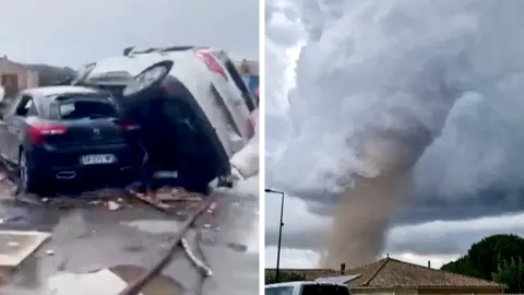 A tornado is seen spinning above a house in the clouds, with two cars on top of each other on the right.