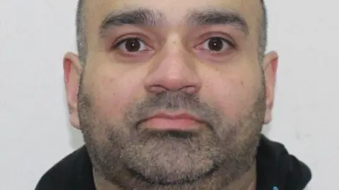 A custody photo man with a shaved head and facial hair, wearing a dark hooded sweatshirt. 