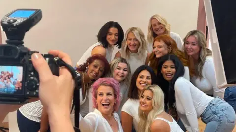 Suministró un grupo de mujeres de diferentes edades y etnias posan para una foto frente a una cámara. Todos visten camisetas blancas y jeans azules.