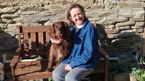 Caroline Roberts Caroline is pictured sat on a brown wooden bench. She is wearing grey trousers and a dark blue denim shirt. She is smiling and looking directly at the camera. Next to her is a brown cocker spaniel. Behind her is a brick wall. Some green plants can be seen in the far right on the floor.