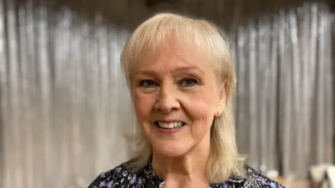 Clare Snow is smiling for the picture. She has blond hair and is in front of a background with silver curtains.