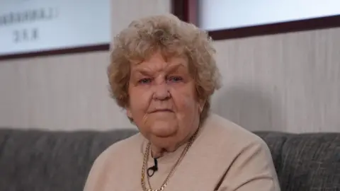 Woman in pale pink sweater wearing a gold chain necklace and short, curly blonde hair, looks at the camera