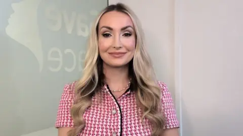 Ashton Collins, who has long blonde hair, looks straight at the camera while smiling. She is wearing a pink and red chequered top, with a white wall and glass panel behind her. 
