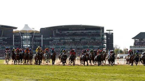 Horses racing at the 2023 Grand National. The stand behind them reads Randox. There is sand coming up from the ground as the horses stampede with jockeys standing up. 