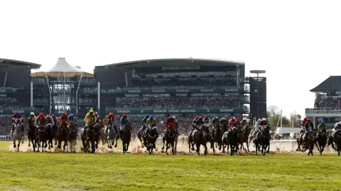 Reuters Horses racing at the 2023 Grand National. The stand behind them reads Randox. There is sand coming up from the ground as the horses stampede with jockeys standing up. 