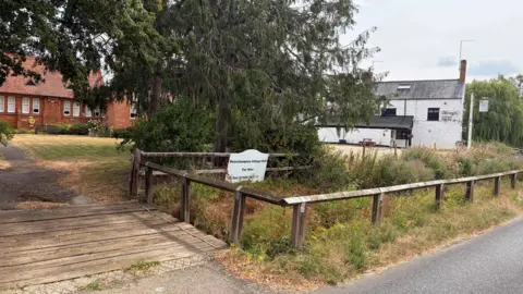 Amy Holmes/BBC Village green with a wooden footbridge, trees, and signs for Beachampton Village Hall, with buildings and a country road in the background.
