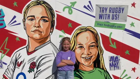 A painted mural depicting England rugby player Sarah Bern and nine year old Ava-Mai smiling and looking at the artist with red and white stripes in the background and colourful arrows and stars on it. The real Ava -Mai stands with her arms folded in front of it, she is smiling at the camera and wearing a purple t shirt and denim shorts