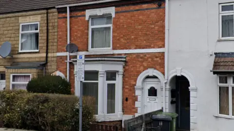 Google A red bricked terraced house is in the middle of the image. The doors and windows are painted white, and a gate is on the outside of the building with a bush. A white parking sign sits in front of the house.