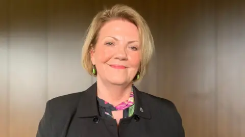 A woman with blonde hair and green earrings in a black jacket with a colourful scarf or blouse underneath