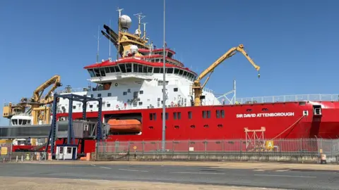 A ship is docked harbourside. It is mainly red-coloured with a white middle deck. 