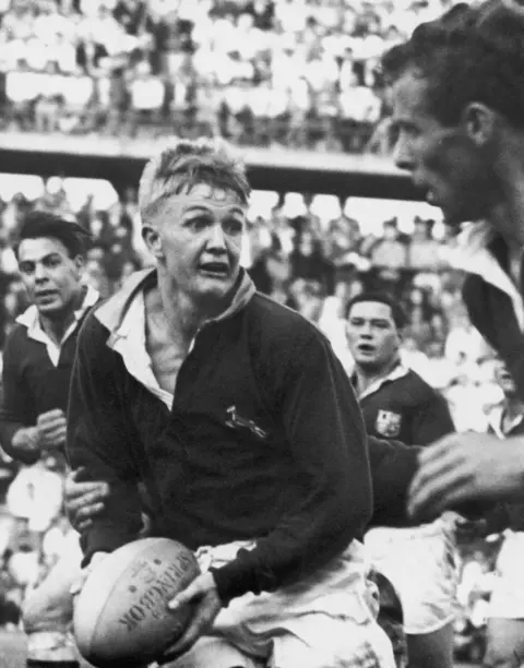 Getty Images A black and white photograph of a rugby match. One man is running with the ball, being pursued by three others. 