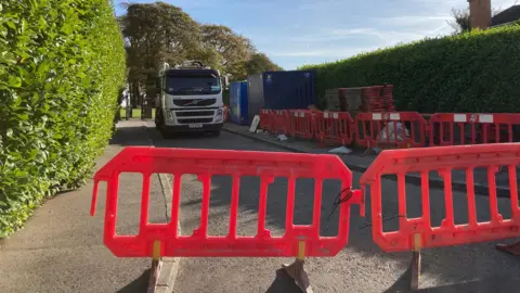 A white truck is parked along the road with orange barriers placed at the end.