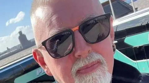 Stuart Burns has grey hair and a grey goatee beard. He wears black sunglasses.