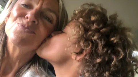 Susie James Susie James and her son who has curly hair and is wearing a grey hoodie. Susie has shoulder-length hair and is wearing a white hoodie. 