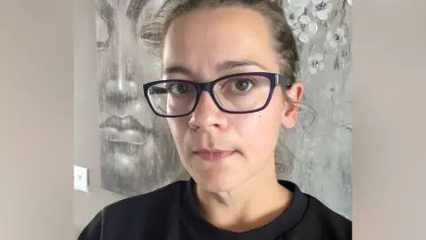 A woman wearing a black and white striped top looks at the camera with a serious expression. She is wearing glasses with dark coloured frames. She has a small silver nose ring. 