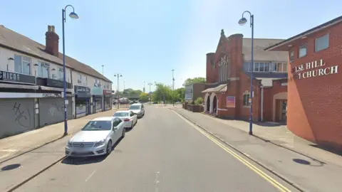 A screenshot of Google street view on a sunny day, showing a road that opens up onto a roundabout in the distance. On the right there is the Gorse Hill Baptist Church, which is a large brick building with stained glass windows. Opposite are several shops with their shutters pulled down and graffiti tags sprayed on them. There are several white cars also parked on this side of the street.