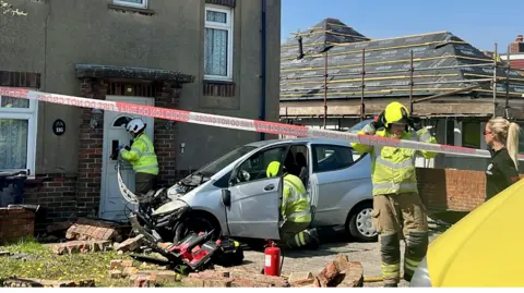 Eddie Mitchell The image shows a car that has crashed into a house; its front is extremely damaged and firefighters can be seen securing the area. A paramedic in a black shirt is also on the scene.