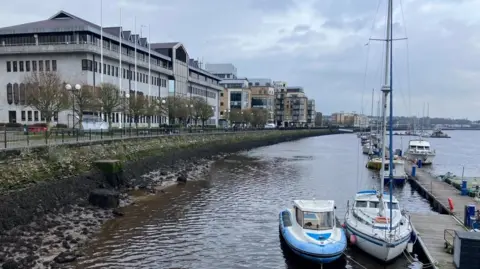A general view of the Foyle riverfront. There is a wooden quay and a small number of boats moored. There are buildings in the background.