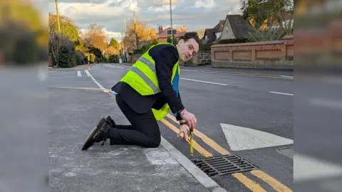 Ted Parton Ted Parton, in a hi vis jacket on Park Lane, measuring the height of the speed table at the edge of the road.