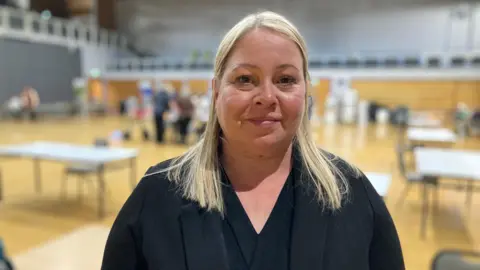 Sarah Sanderson / BBC A head and shoulders shot of Emma Clough smiling at the camera while stood in a hall. She has long, straight, blonde hair and is swearing a black blazer and a black blouse. 