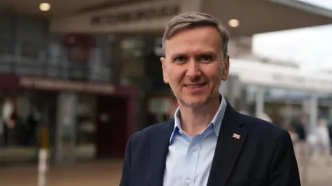 Martin Giles/BBC Andrew Pakes stares at the camera. He wears a light blue shirt with a dark jacket. A small union flag badge is on his left hand side. 