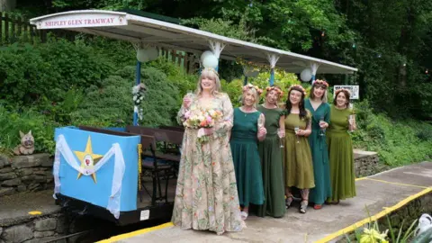 A bride in a floral white dress stands next to a tram with a line of bridesmaids in green dresses behind her