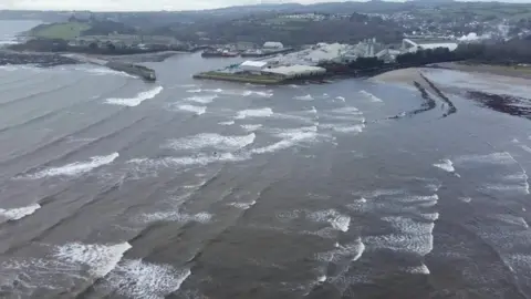 An aerial view of a bay with a harbour in the distance and a beach.