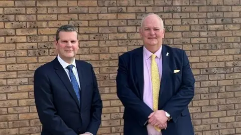 Humberside Police Chris Todd stands next to Jonathan Evison. Mr Todd is wearing a dark blue suit, a white shirt and a blue tie. Mr Evison is wearing a dark suit, a pink shirt and a yellow tie. They are standing in front of a brick wall.