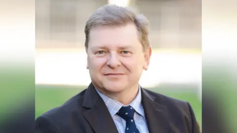 Royal School Dungannon Dr David Burnett. He has short silvery brown hair, wearing a dark blazer, light blue check shirt and navy tie with white dots.