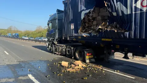 A lorry with the side of its trailer ripped. Boxes of wine are visible, with some smashed and crumpled in the road. The carriageway is clear of traffic, though there is a queue on the other side.