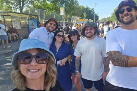 Manchester University NHS Foundation Trust Woman with long blonde hair, blue hat and sunglasses, man with beard, brown hat, white top and sunglasses, woman with long dark hair, blue dress and sunglasses, woman with dark hat, blue dress and sunglasses, man with beard, dark hat and light top with dark shorts and a man with beard, dark hat, sunglasses and white top stood outdoors.