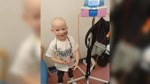 Jaxson in a hospital room with no hair, a tube coming out of his nose, and attached to a small machine on a stand. He is wearing a t-shirt saying 'Fighter'