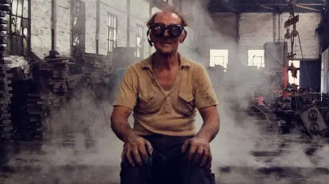 Diana Grandi Colour image of a man with messy hair and wearing large protective goggles over his eyes. He is wearing a beige t-shirt and black trousers and is seated facing the camera with his hands on his knees. Behind him there are pulleys and machines which can be seen through wisps of smoke.