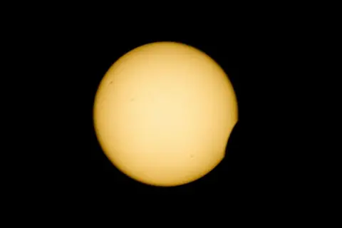 EPA/Shutterstock A yellow circle – the Sun – sits against a black sky. There is a small crescent “bite” of black removed from the lower right corner as the Moon moves across it 