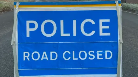 BBC Stock image of a blue sign in the middle of a road. It reads 'Police Road Closed'.