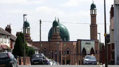 A street view of Makkah Masjid in Leeds. It has a large green dome and a striped brick design.