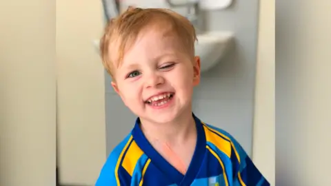 Young boy with blue eyes and blonde hair smiles in sports shirt. He is in a cream-walled room and has one eye shut.