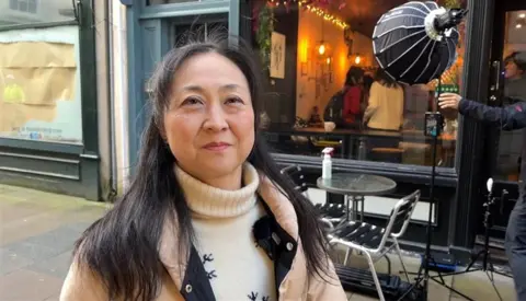 BBC Japanese woman stood outside Bean Jar cafe in Guernsey. There is a film crew light just outside the cafe and a small table. 