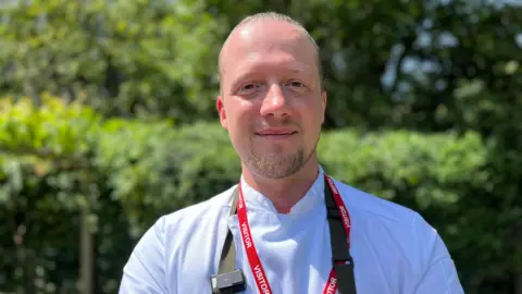Luke is smiling at the camera. His hair is pulled back on the top of his head. Behind him, are the bushes surrounding the allotment. He is wearing a white chef top with a green apron. 