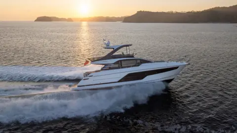 A small white yacht makes its way across water, with a large wash behind it